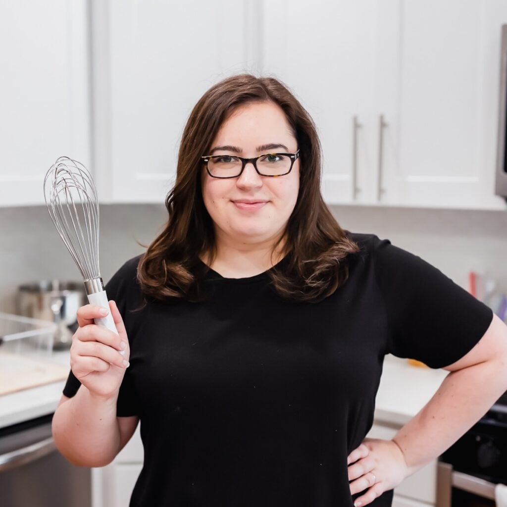 A woman with glasses and dark hair stands in a kitchen, wearing a black shirt and holding a whisk in her right hand. She faces the camera and smiles slightly. White cabinets and kitchen items are visible in the background.