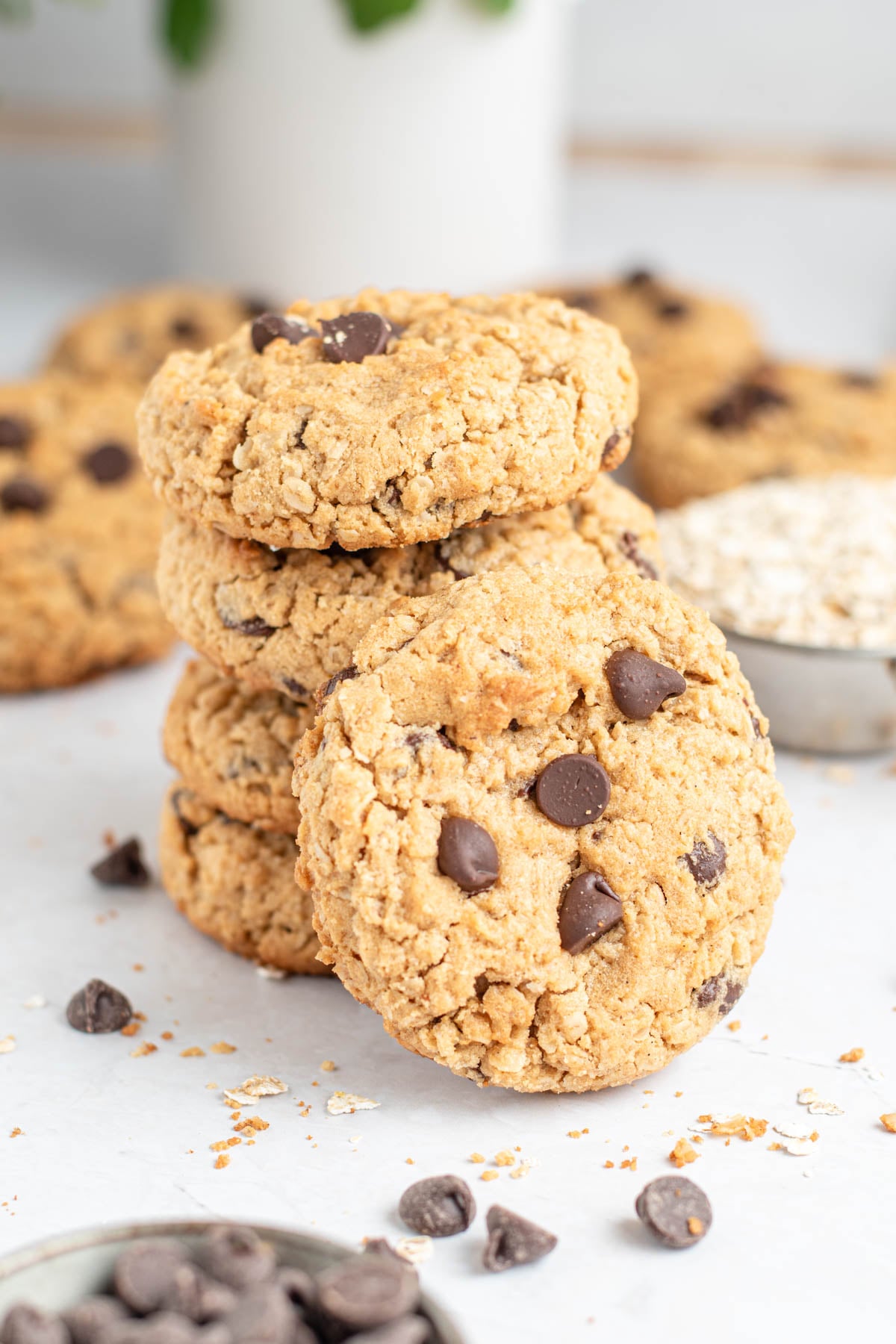 A stack of four oatmeal chocolate chip cookies is shown on a white surface, reminiscent of classic peanut butter cookies, with extra cookies, chocolate chips, and a small bowl of oats in the background.