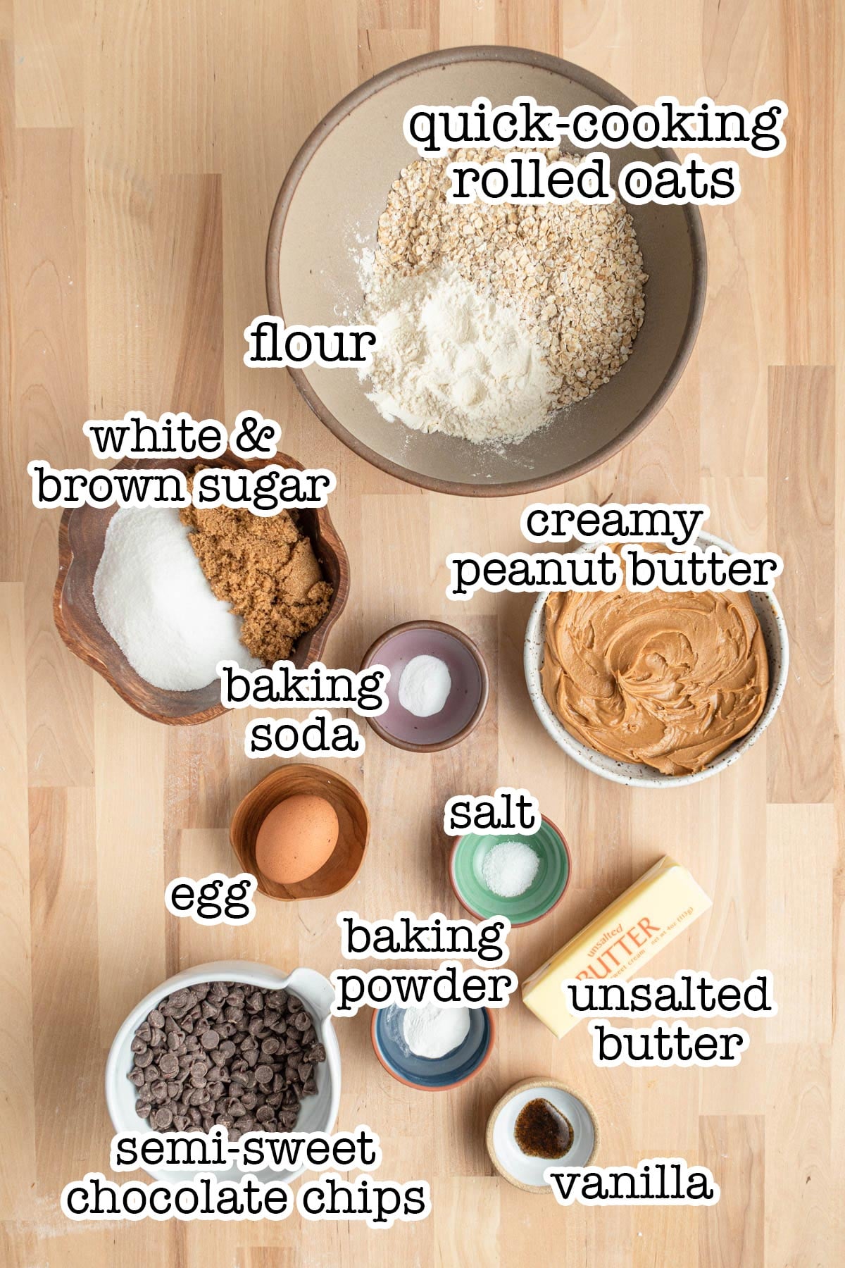 A top-down view of labeled baking ingredients for oatmeal peanut butter chocolate chip cookies on a wooden surface: quick-cooking rolled oats, flour, peanut butter, sugars, baking soda, salt, egg, baking powder, unsalted butter, vanilla, and chocolate chips.