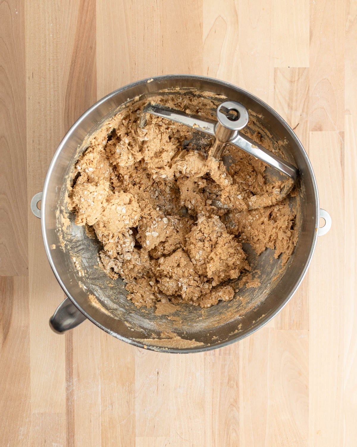 A metal mixing bowl containing thick peanut butter cookie dough and a paddle attachment sits on a light wooden surface. Some dough and flour are visible on the sides of the bowl.