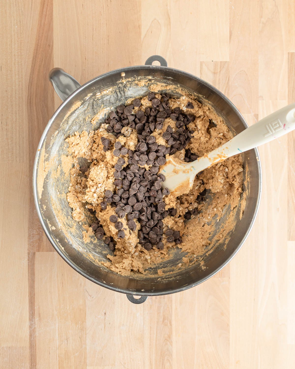 A metal mixing bowl on a wooden surface contains peanut butter cookie dough with chocolate chips being mixed in with a white spatula.