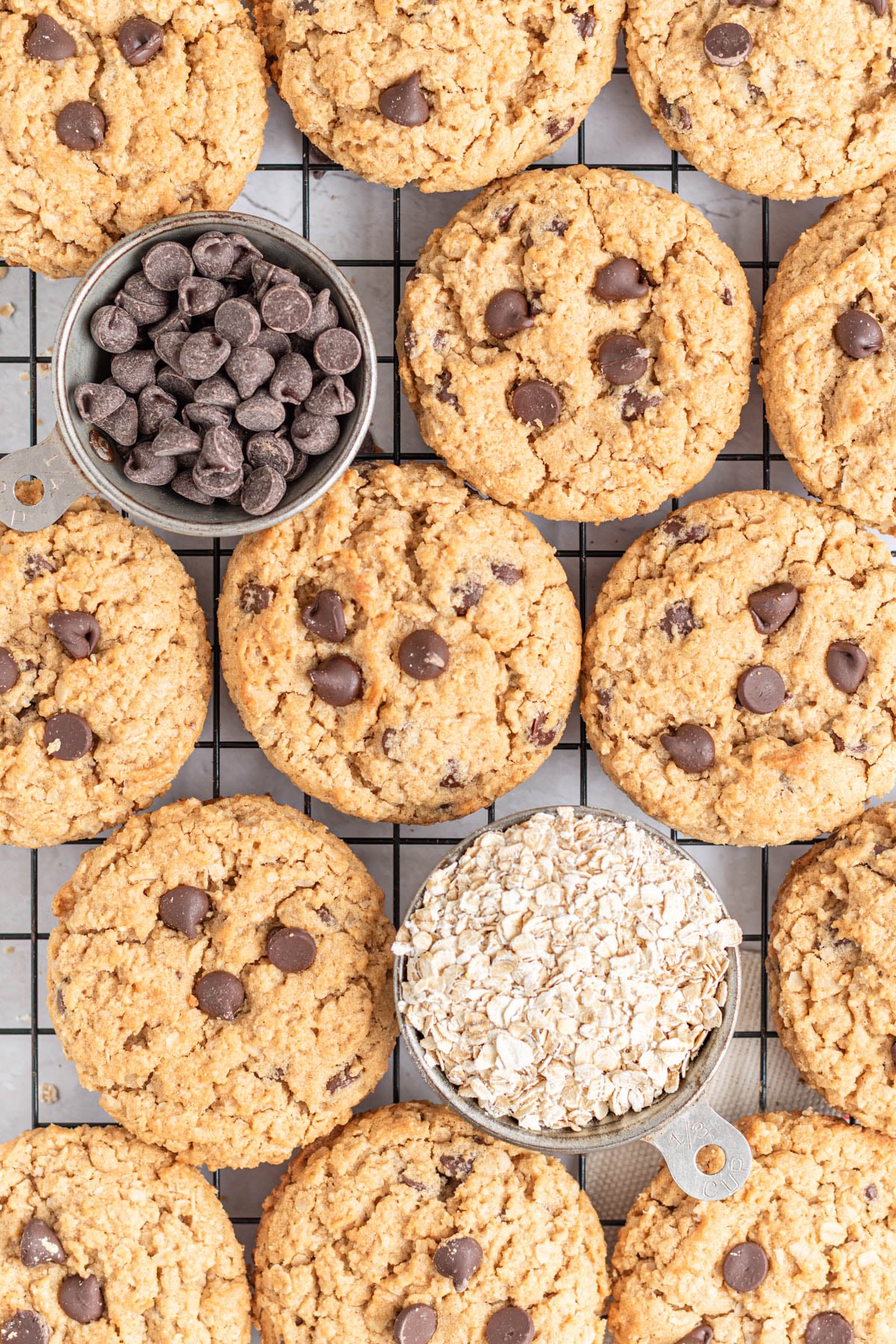 Chocolate chip oatmeal cookies are arranged on a cooling rack. Among the cookies—perfect for fans of peanut butter cookies—are two metal measuring cups, one filled with chocolate chips and the other with rolled oats.