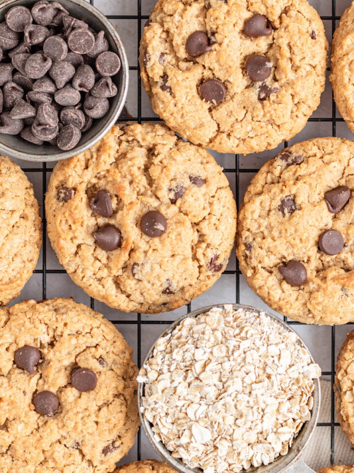 Chocolate chip oatmeal cookies are arranged on a cooling rack with two small bowls—one filled with chocolate chips and the other with rolled oats—giving a cozy, homemade vibe perfect for fans of classic treats like peanut butter cookies.