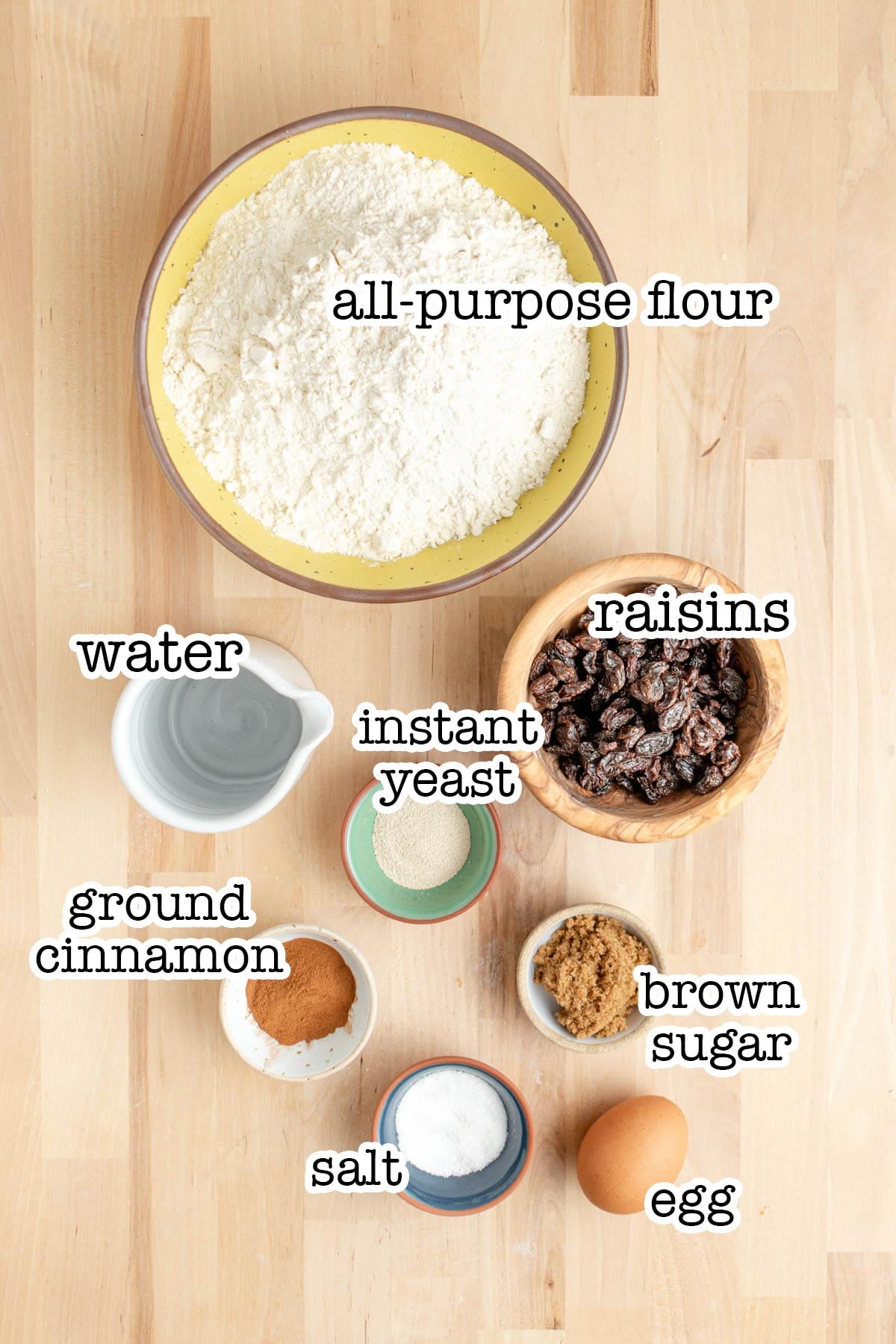 A top-down view of labeled bowls containing all-purpose flour, raisins, brown sugar, water, instant yeast, ground cinnamon, salt, and an egg—ingredients arranged on a light wooden surface for homemade cinnamon raisin bagels.