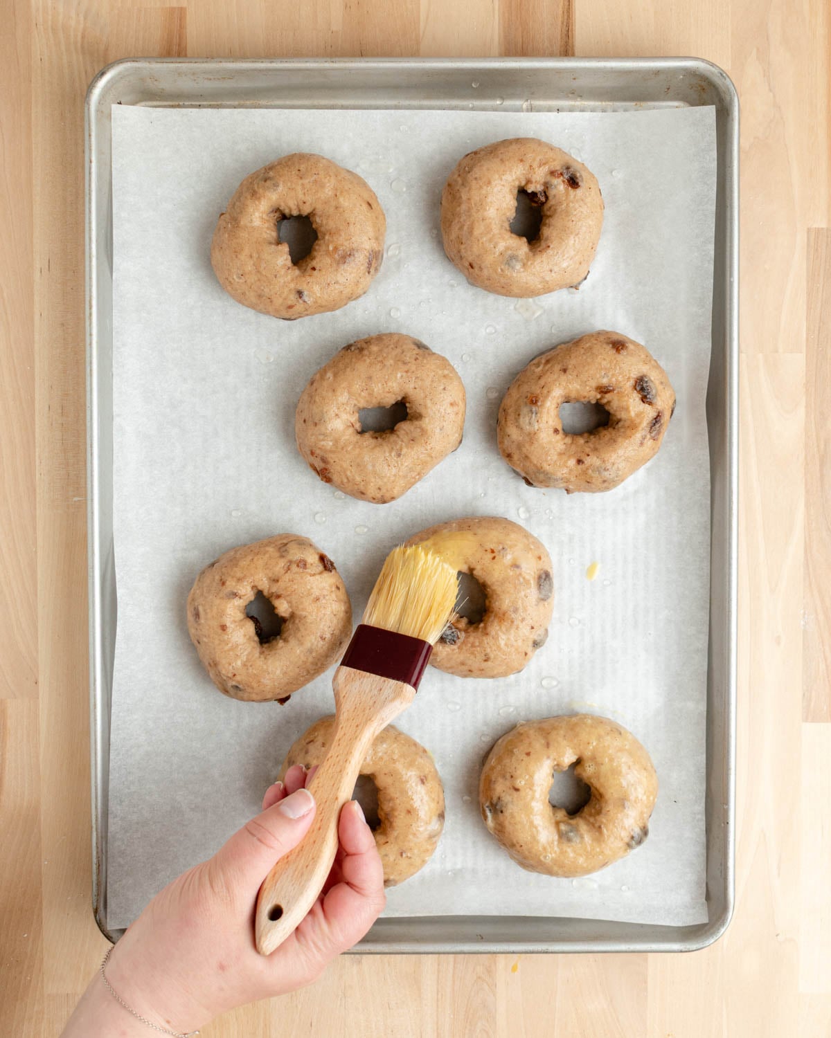 A hand holds a pastry brush, applying egg wash to unbaked, round raisin bagels, arranged on a parchment-lined baking sheet.