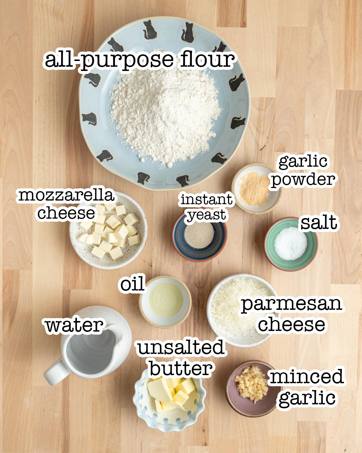 A bowl of flour sits on a wooden table, along with other labeled ingredients: mozzarella cheese, oil, water, unsalted butter, minced garlic, parmesan cheese, salt, instant yeast, and garlic powder.