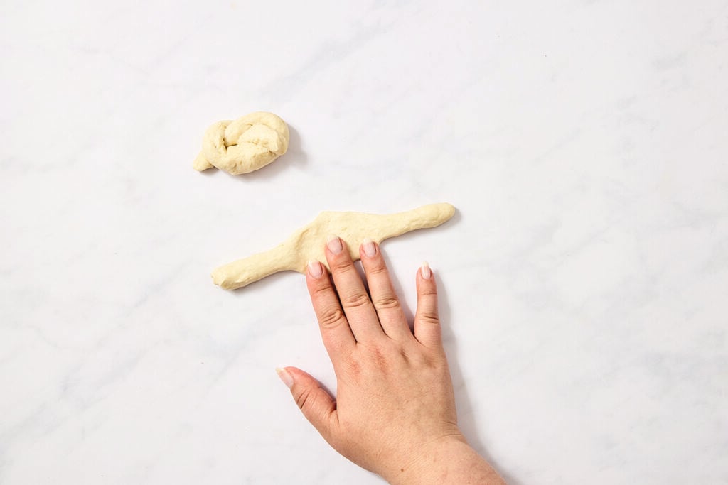 A hand presses on a piece of dough, rolling it into a rope shape on a white surface. A small, twisted piece of dough sits above the hand.