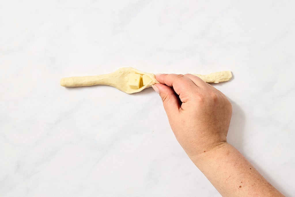 A hand folds a piece of dough around cubes of butter on a white surface. The dough is stretched out, and the butter is placed in the center before being wrapped.