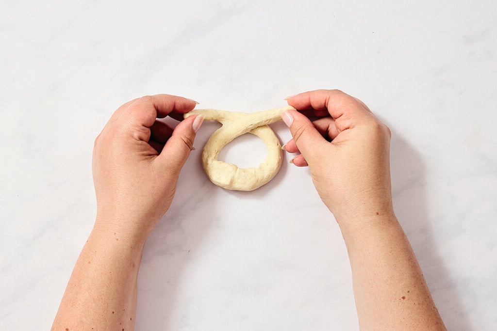 Two hands are shaping a piece of dough into a ring on a white surface.
