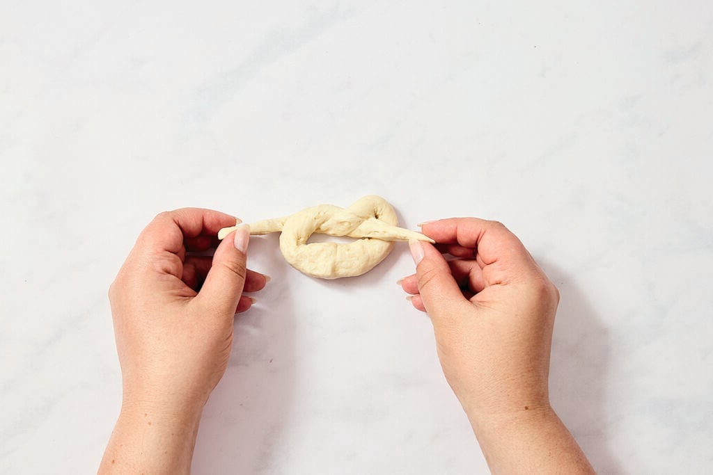 Two hands are shaping a piece of dough into a pretzel shape on a white, lightly textured surface.