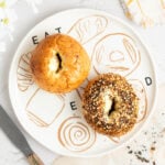 A plate with “EAT BREAD” written on it holds two bagels; one is plain and the other is topped with everything seasoning. A serrated knife and a napkin are nearby on a white surface.