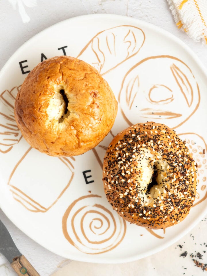 A plate with “EAT BREAD” written on it holds two bagels; one is plain and the other is topped with everything seasoning. A serrated knife and a napkin are nearby on a white surface.