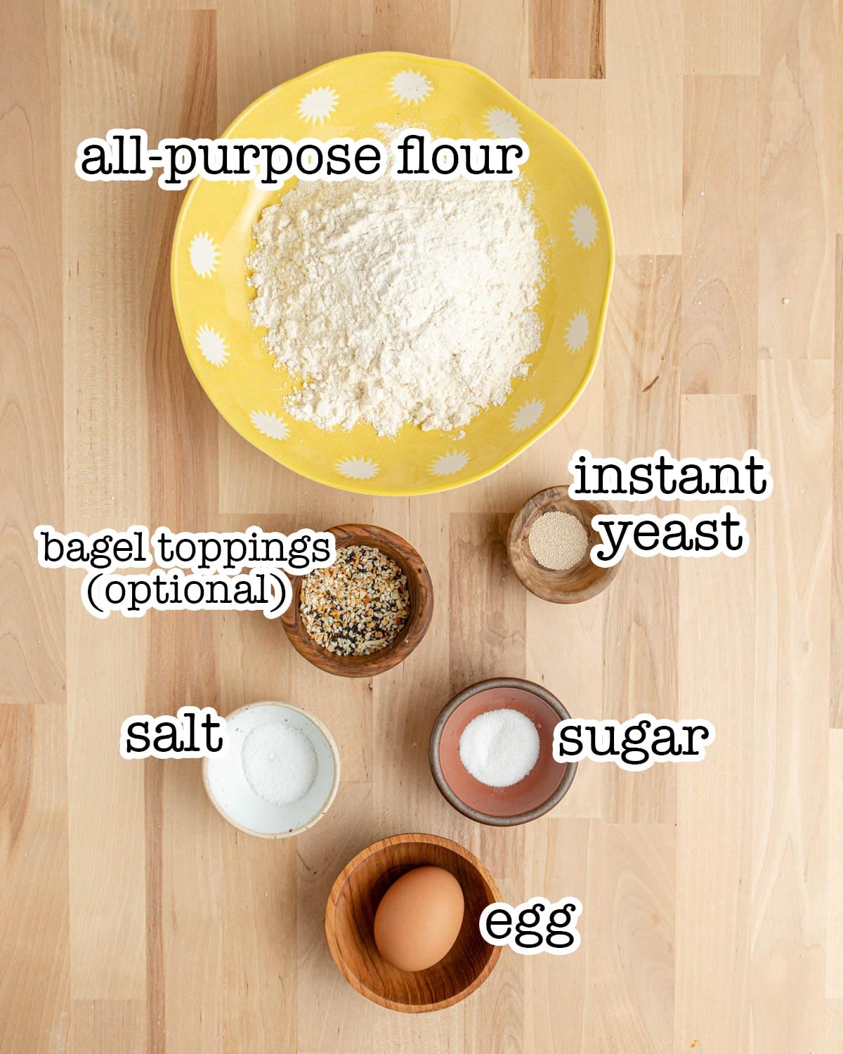 A yellow bowl with flour, and small bowls containing instant yeast, salt, sugar, an egg, and bagel toppings on a wooden surface. Each ingredient is labeled with text.