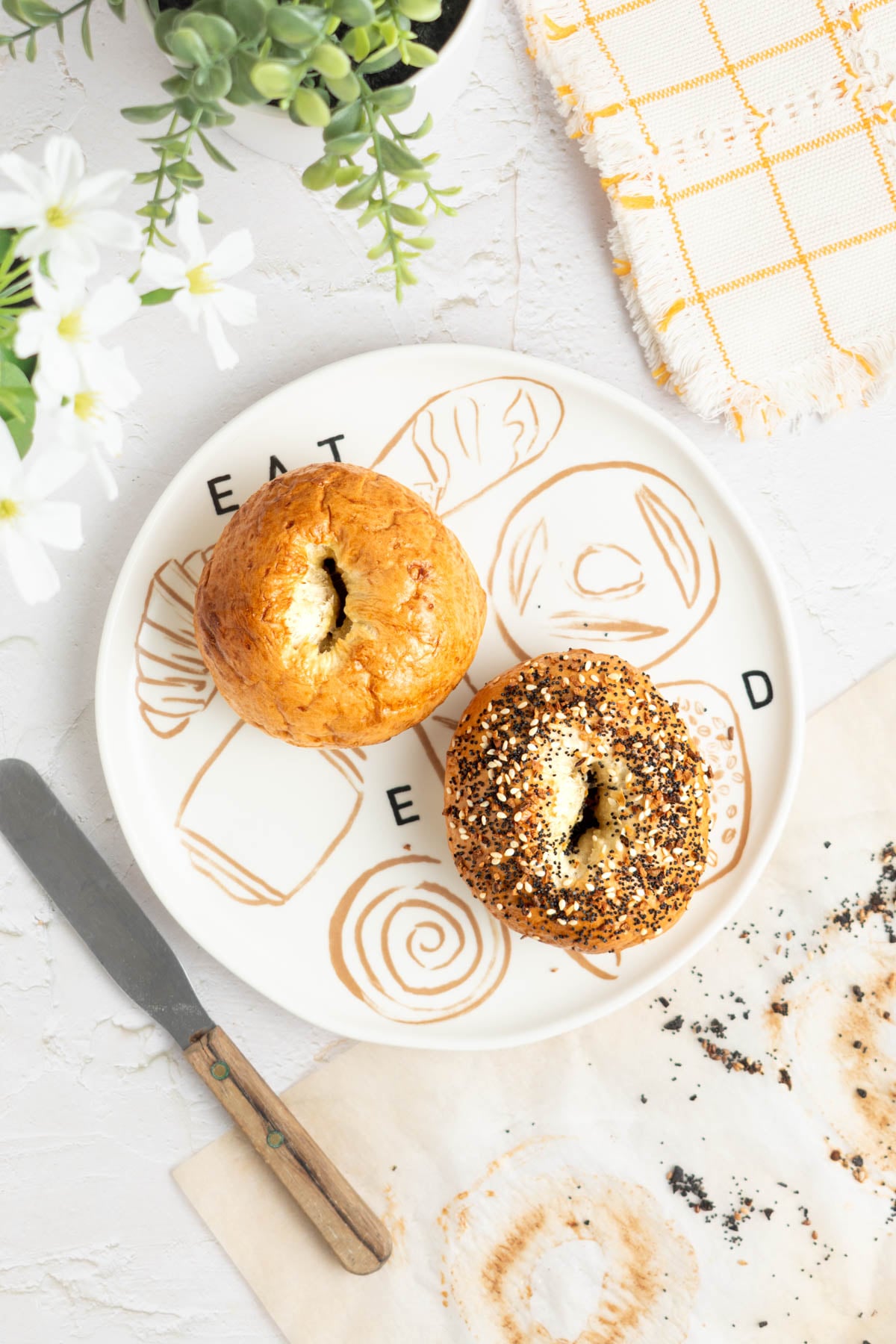 A plate with “EAT” "BREAD" printed on it holds two bagels, one plain and one covered in everything seasoning. A knife, napkin, parchment with crumbs, and a plant are arranged on a white surface nearby.