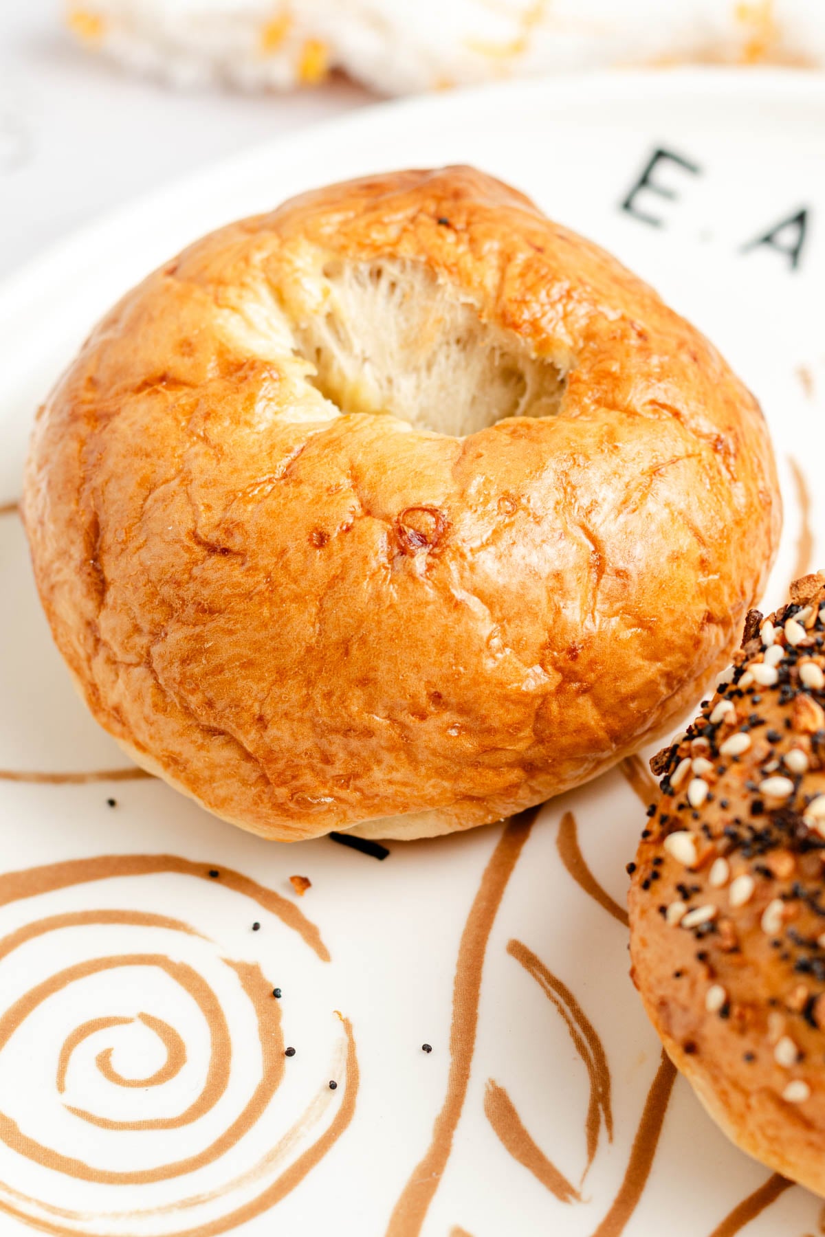 A plain bagel with a shiny, golden-brown crust sits on a white plate with tan swirl designs. A partially visible everything bagel is next to it.