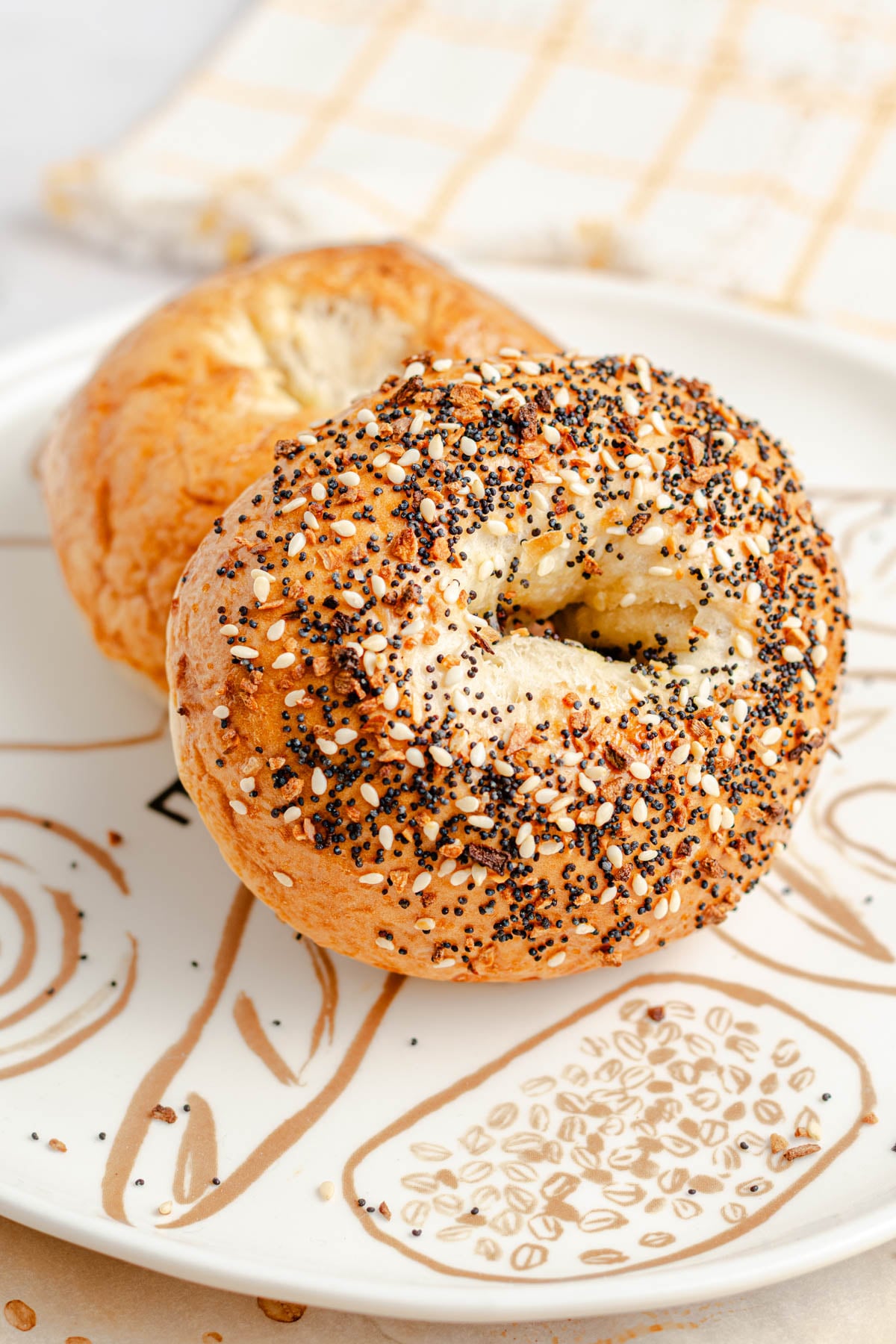 Two bagels with a golden-brown crust and a topping of mixed seeds are placed on a white plate with a beige pattern. A beige and white checkered cloth is in the background.