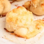 A close-up of a baked garlic knot topped with melted, golden-brown cheese on a sheet of parchment paper. Other cheese-covered knots are visible in the blurred background.