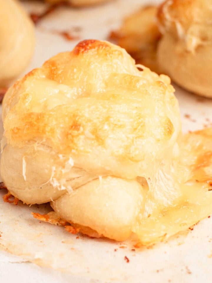 A close-up of a baked garlic knot topped with melted, golden-brown cheese on a sheet of parchment paper. Other cheese-covered knots are visible in the blurred background.
