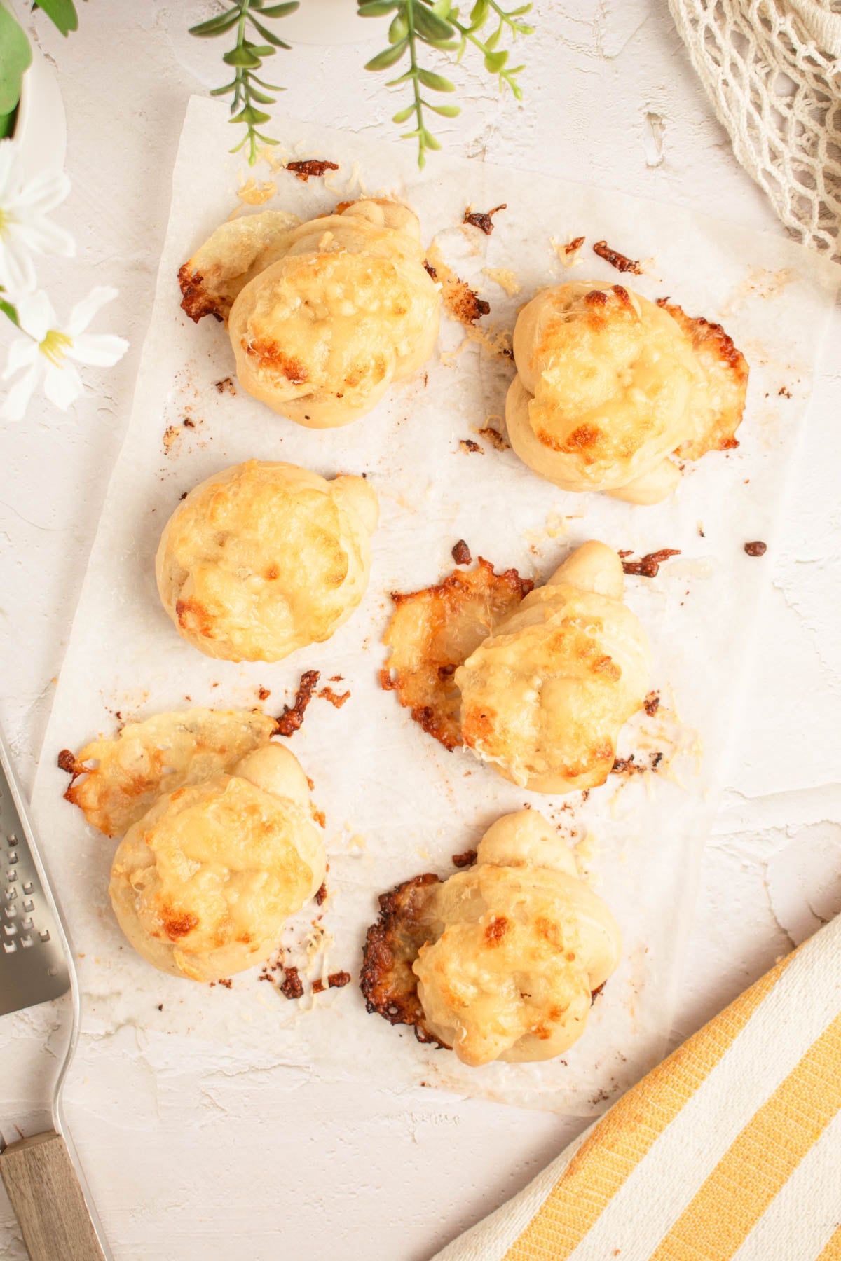 Six baked cheese-covered bread knots are arranged on a piece of parchment paper. Some melted cheese has spread onto the paper. A metal spatula, flowers, and a striped towel are visible around the edges.