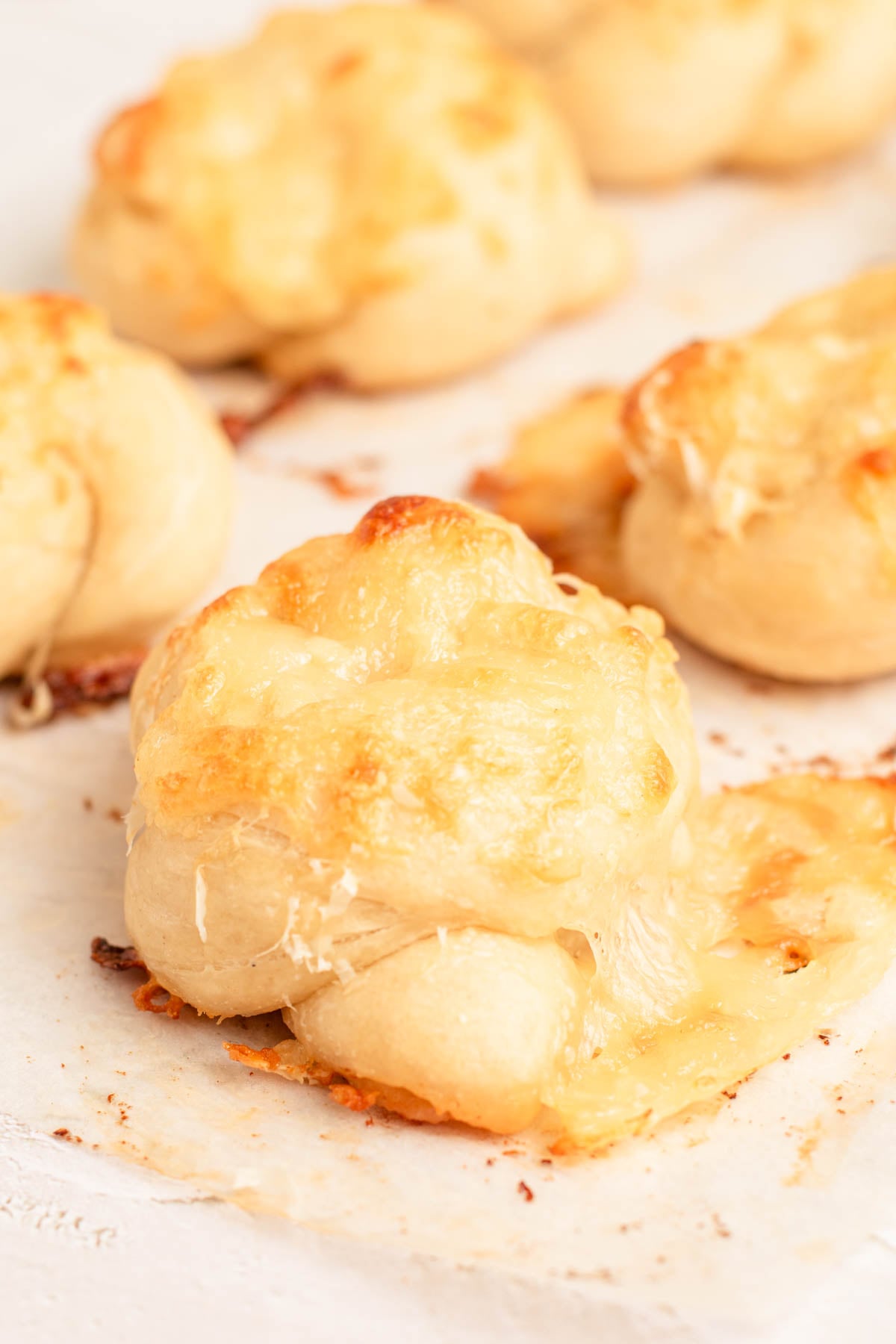 Baked garlic knots topped with melted cheese on a sheet of parchment paper. The cheese is slightly browned on top, and some has melted onto the parchment.