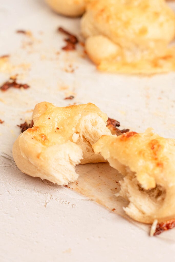 A close-up of a baked cheesy garlic knot torn in half, showing its soft interior and melted cheese on top. The bread sits on a light surface with scattered crumbs and bits of cheese.