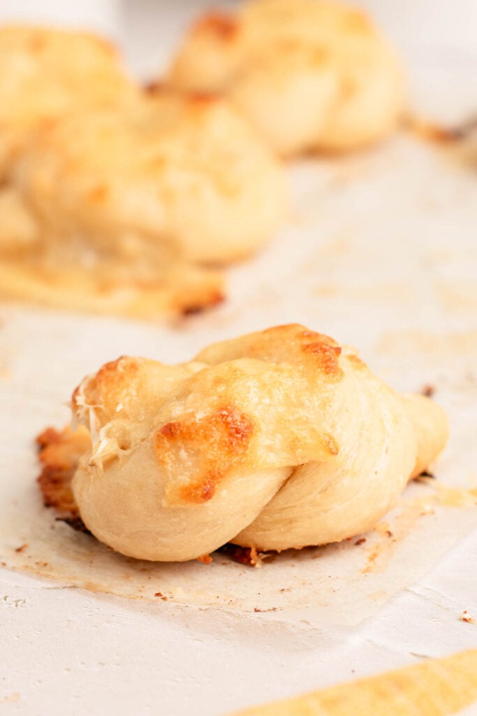A close-up of a baked garlic knot topped with melted cheese, resting on a parchment-lined baking sheet. Other garlic knots are blurred in the background.