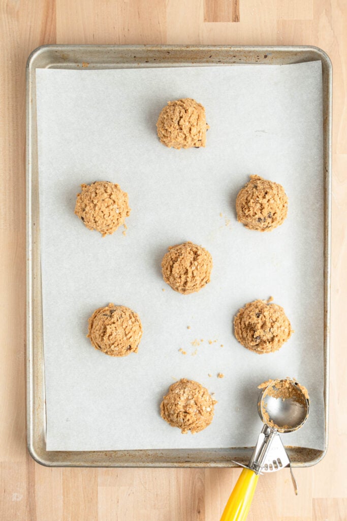 A baking sheet lined with parchment paper holds seven scoops of oatmeal peanut butter cookie dough, arranged in rows. A metal cookie scoop with a yellow handle rests on the lower right corner of the tray.