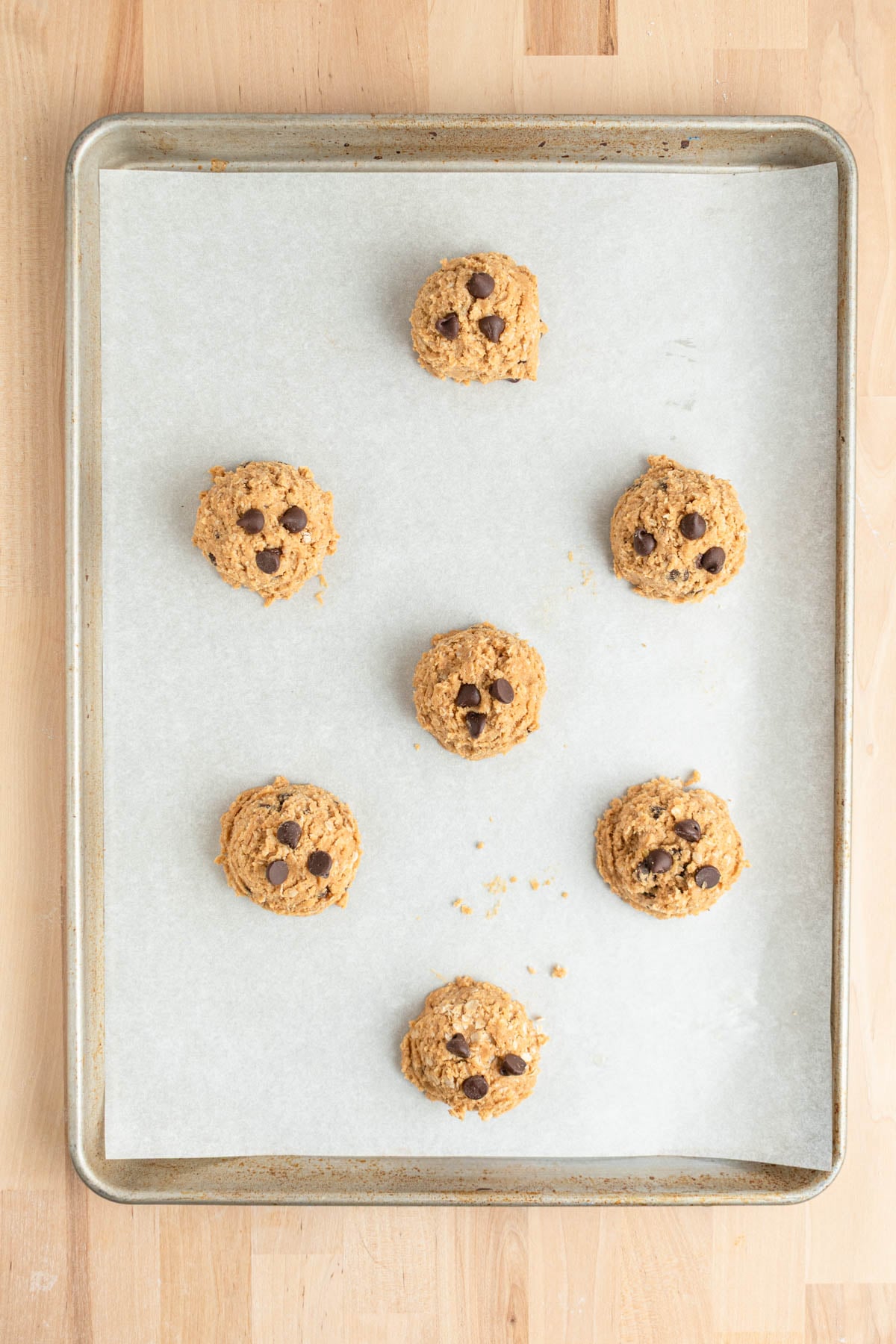 A baking sheet lined with parchment paper holds seven unbaked oatmeal peanut butter cookie dough balls each topped with 3-4 chocolate chips, spaced apart on a wooden surface.