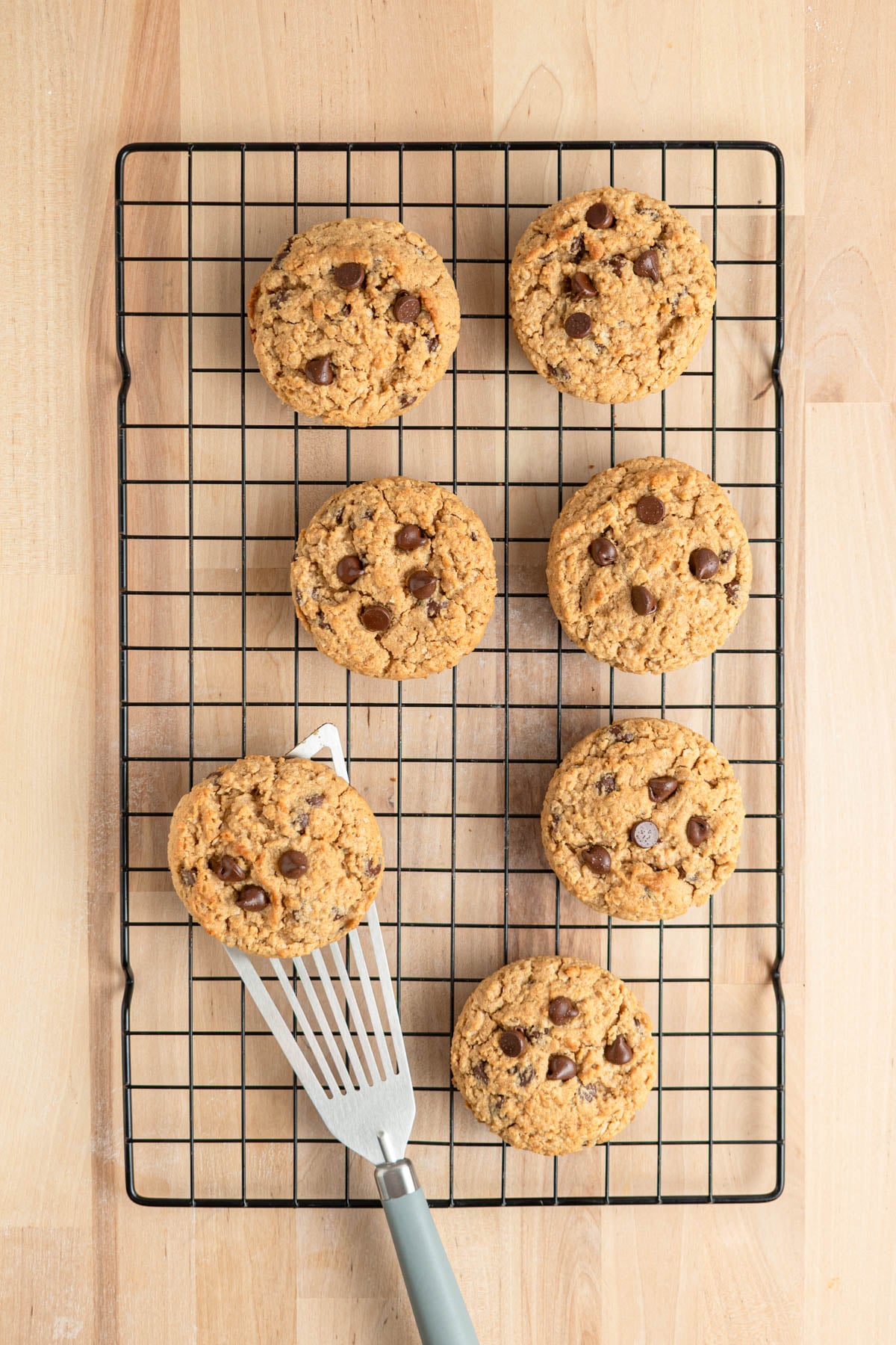 Seven peanut butter cookies are on a black wire cooling rack over a light wood surface. One cookie is being lifted off the rack with a metal fish spatula that has a gray handle.