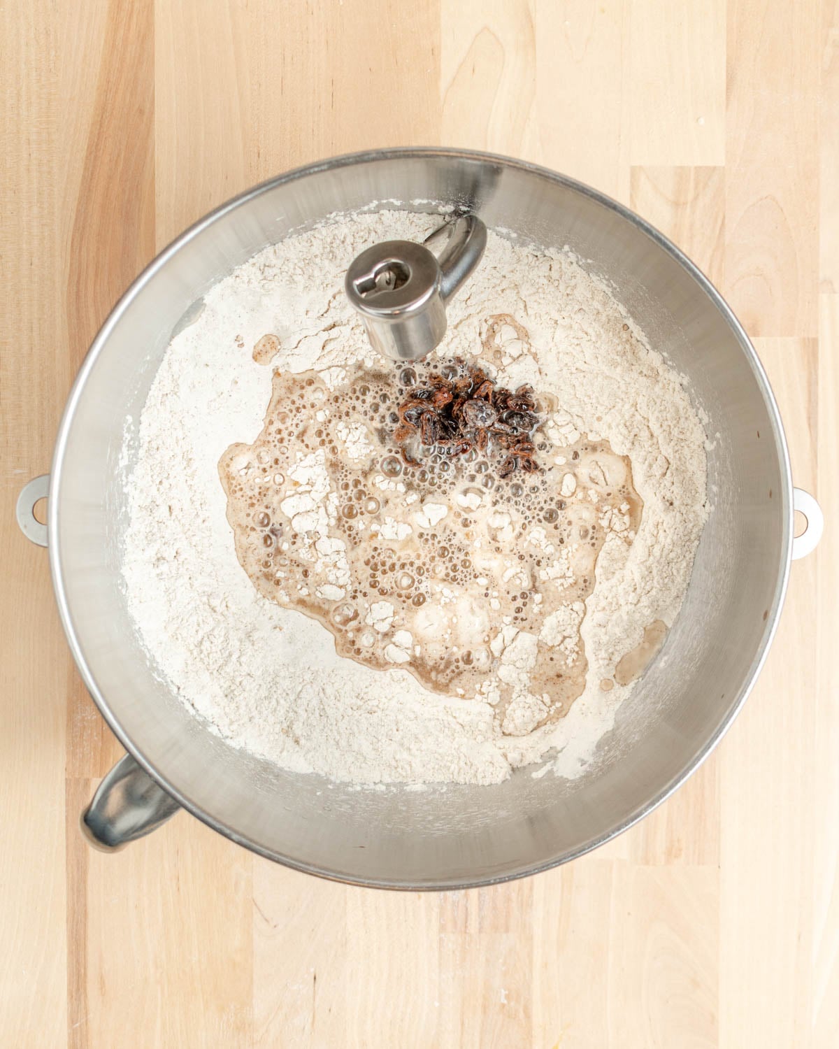 A metal mixer bowl on a wooden surface contains flour, yeast, water, spices—classic ingredients for making raisin bagels—prepared for mixing with an electric dough hook attachment.