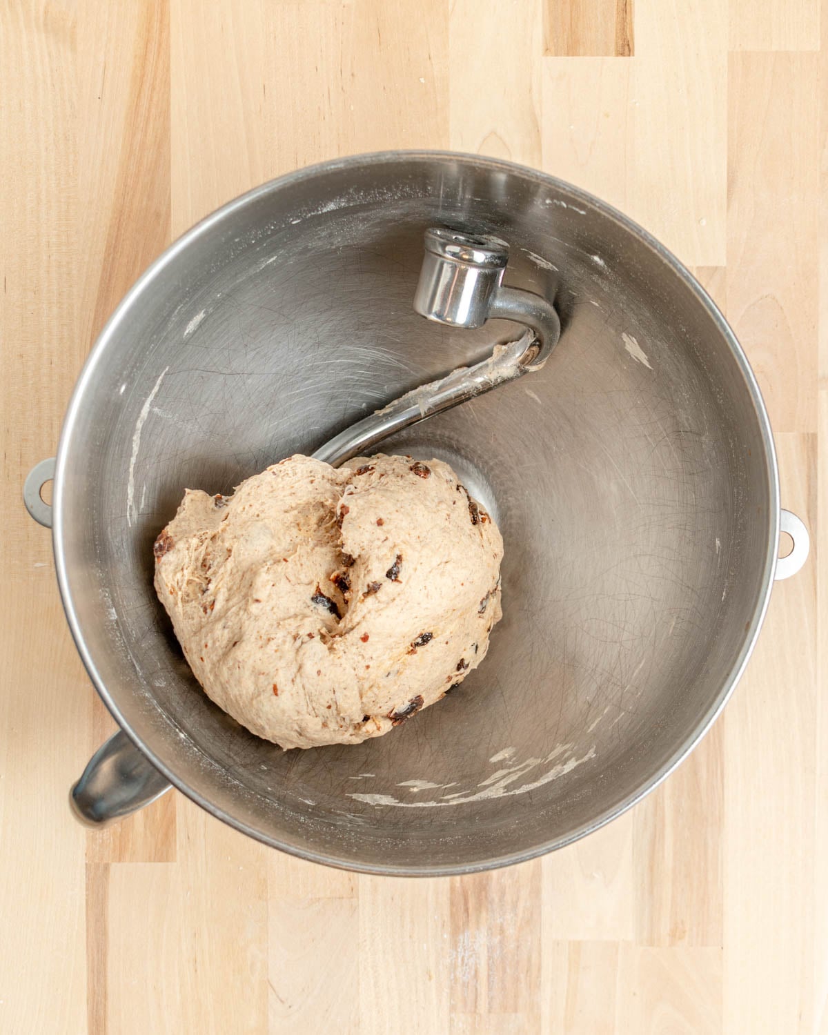 A ball of bagel dough with visible raisins sits in a metal stand mixer bowl with a dough hook attached, on a light wooden surface.