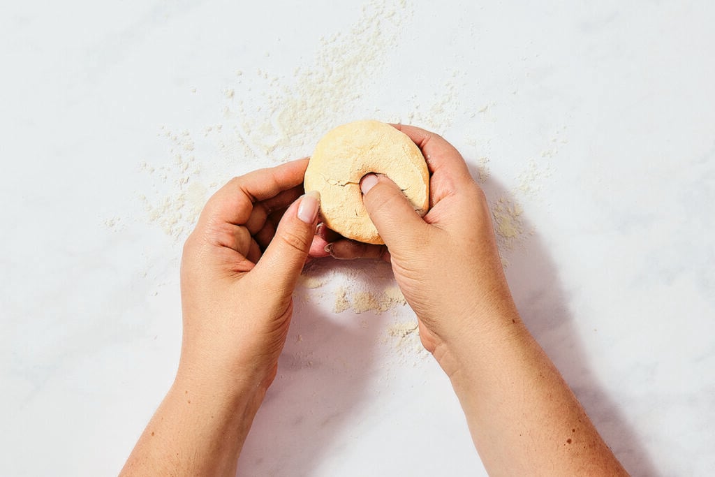 Two hands are shaping a piece of dough into a round form on a white, floured surface—crafting small batch bagels as the thumbs create an indentation in the center of the dough.
