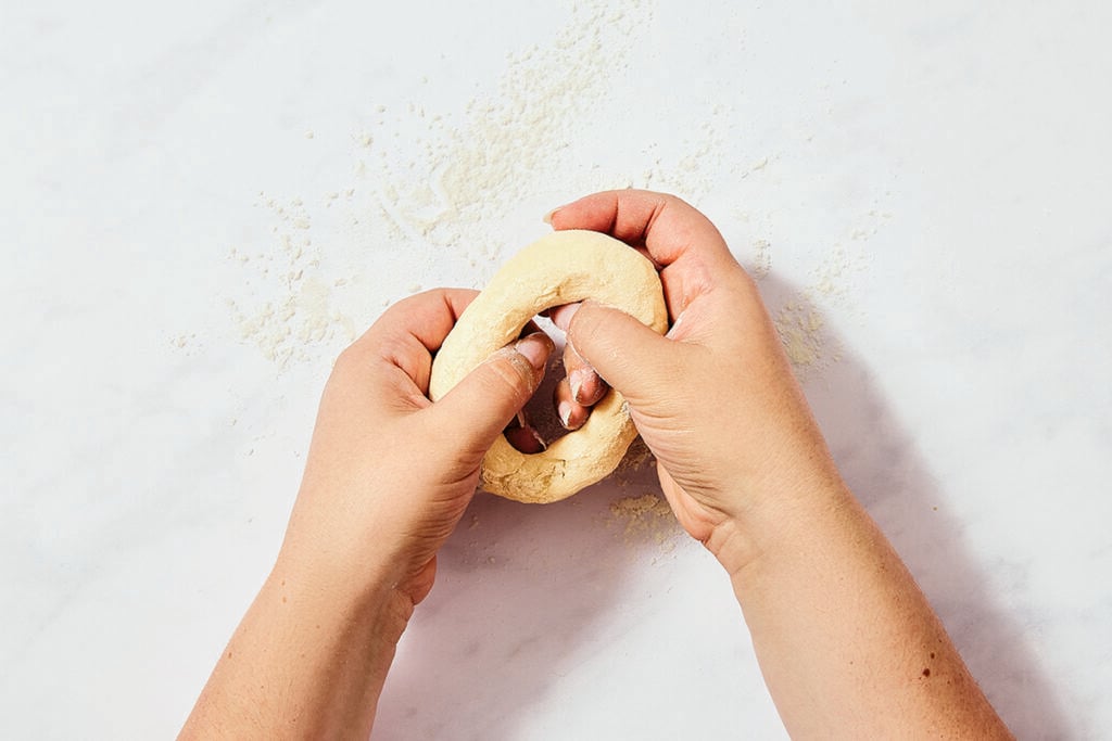 Two hands are stretching a piece of dough into a ring on a lightly floured white surface, preparing small batch bagels.