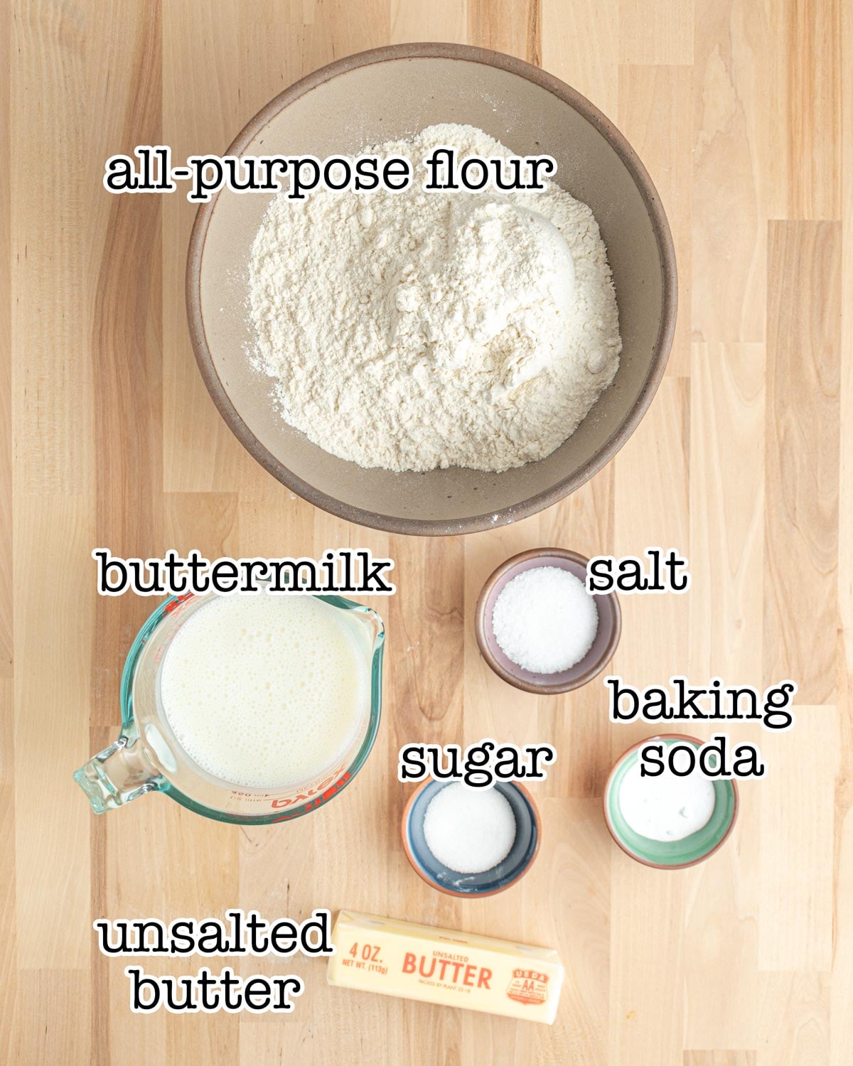 A bowl of all-purpose flour, a glass measuring cup of buttermilk, a stick of unsalted butter, and small bowls of salt, sugar, and baking soda are arranged on a wooden surface with labels for each ingredient.