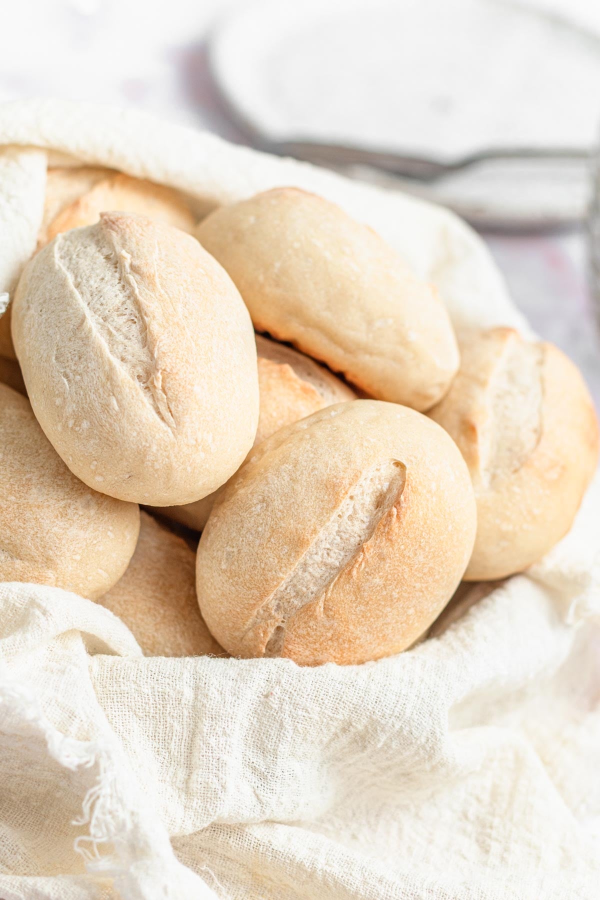 A basket lined with a white cloth holds several small, round, crusty bread rolls with a slit down the center. The background is light and out of focus, highlighting the fresh bread.