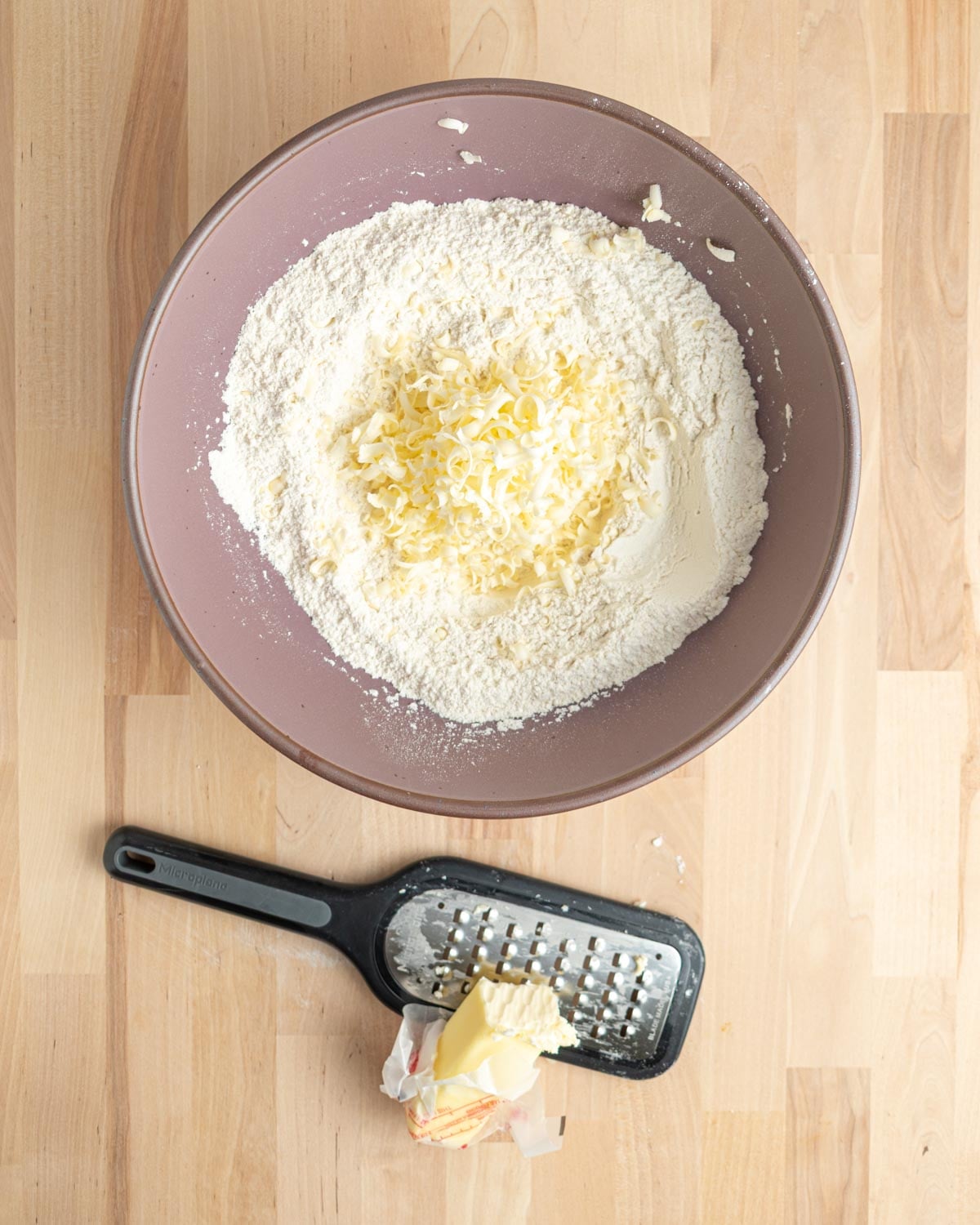 A mixing bowl filled with flour and grated butter sits on a light wooden surface. Below the bowl, a coarse paddle grater with a partially grated stick of butter rests on the table.