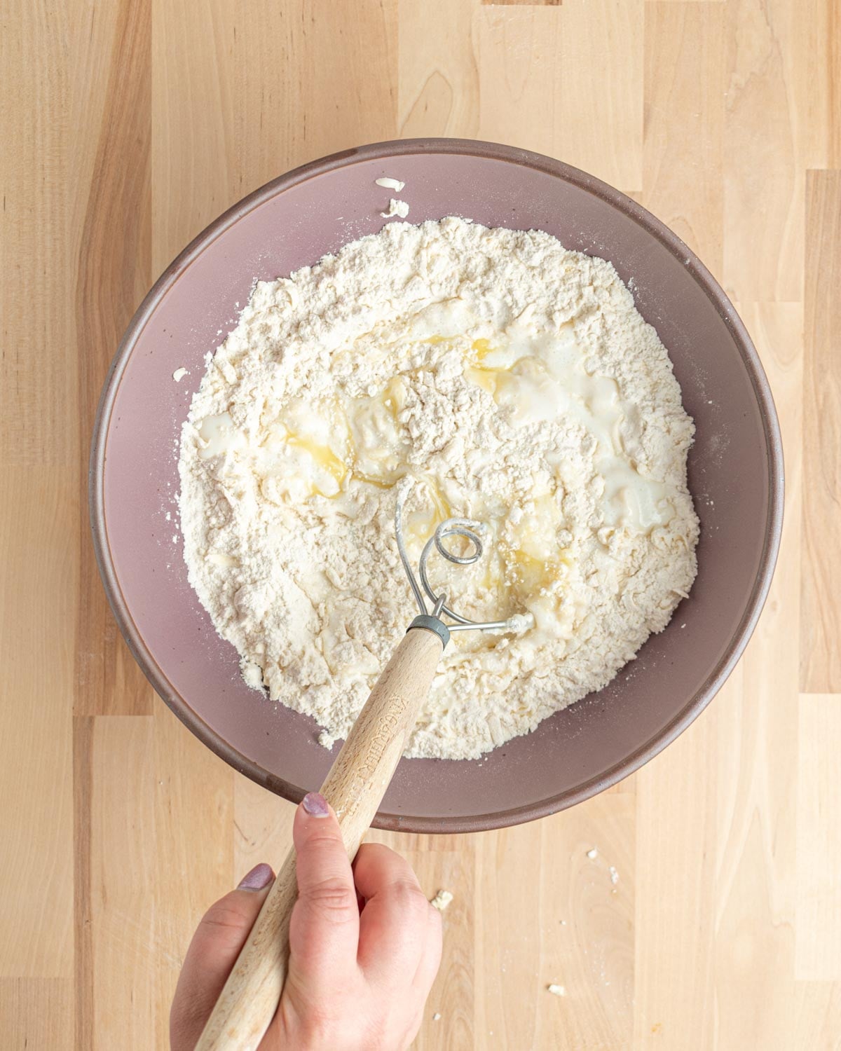 A hand holds a dough whisk in a bowl of flour mixture with buttermilk and melted butter on top, placed on a light wooden surface.