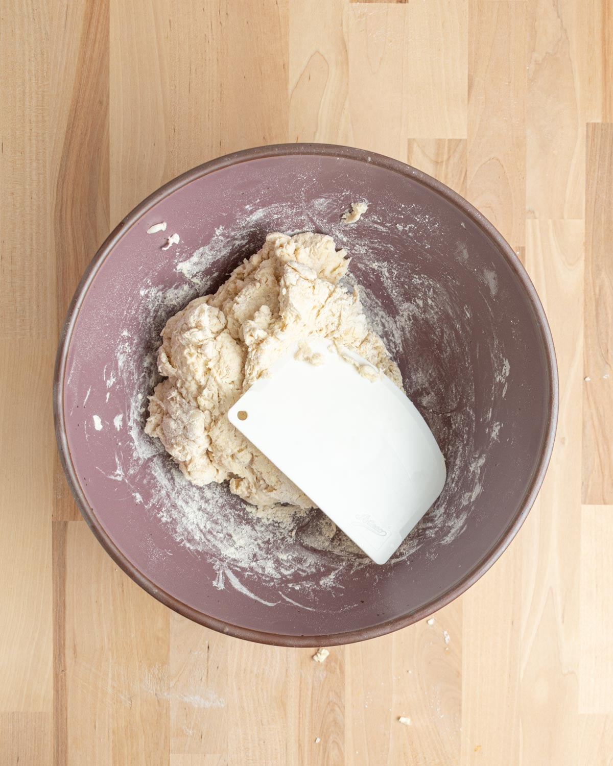 A mixing bowl containing partially kneaded soda bread dough and a white plastic dough scraper, placed on a light wooden surface.