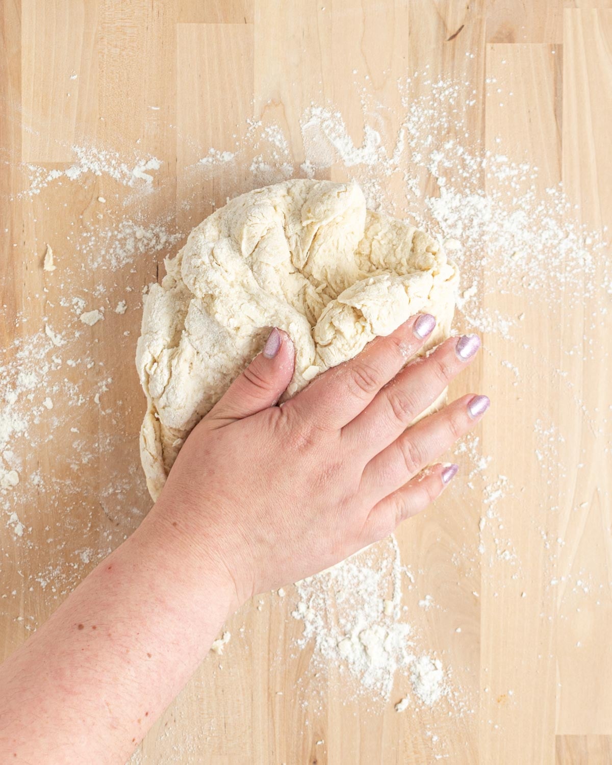 A hand presses down on a mound of dough on a floured wooden surface, with flour scattered around and visible on the persons fingers.