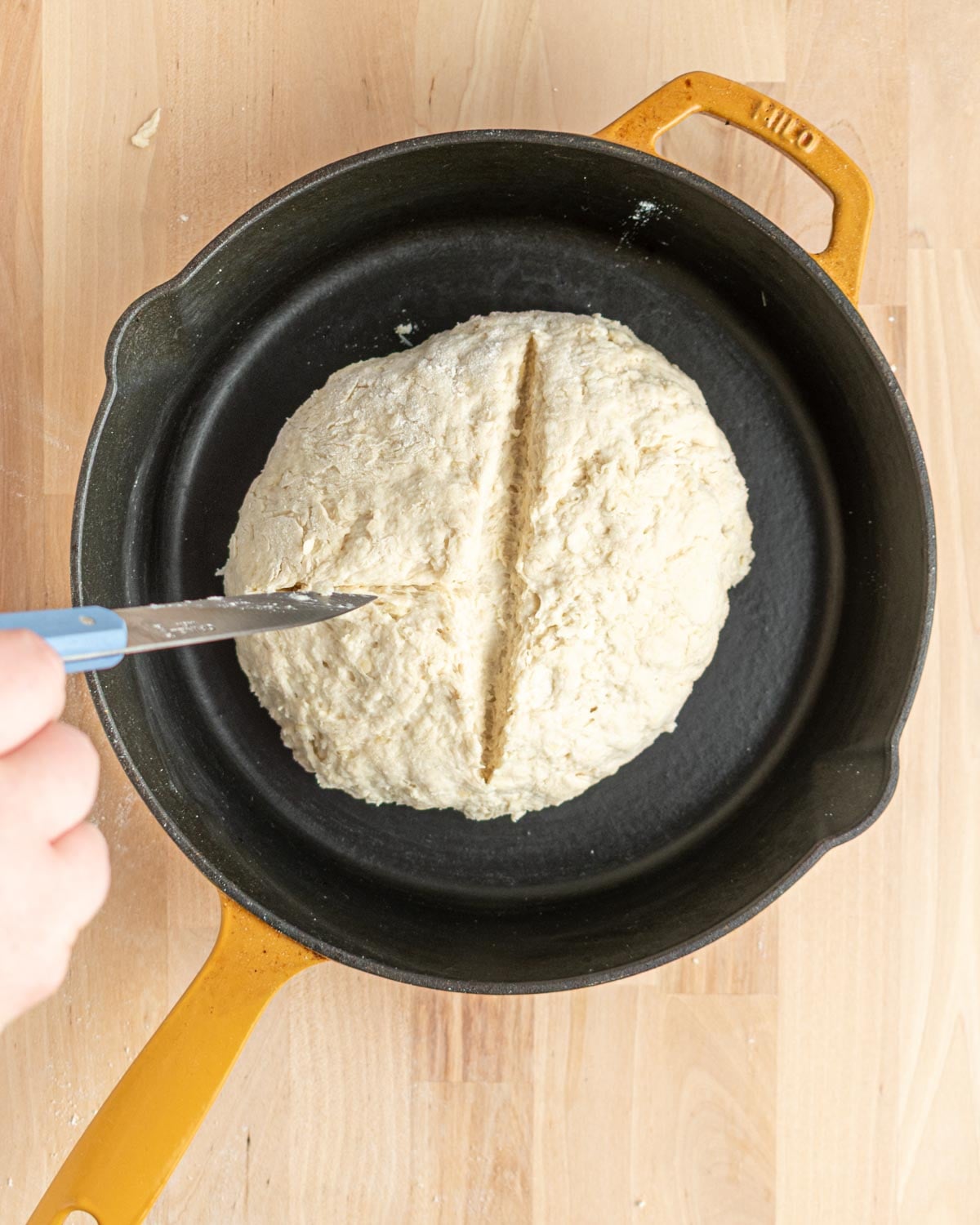 A hand uses a knife to score a line into the top of a round ball of raw dough placed in a black cast iron skillet with a yellow handle, on a light wooden surface.