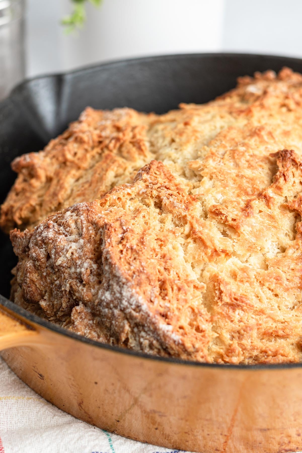 A close-up of a freshly baked loaf of soda bread with a golden-brown, rough crust, sitting in a yellow cast iron skillet. The bread appears rustic and hearty.