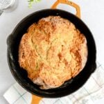 A round loaf of golden-brown soda bread sits in a black cast iron skillet with a tan handle, placed on a white and blue checkered towel, on a light-colored surface.