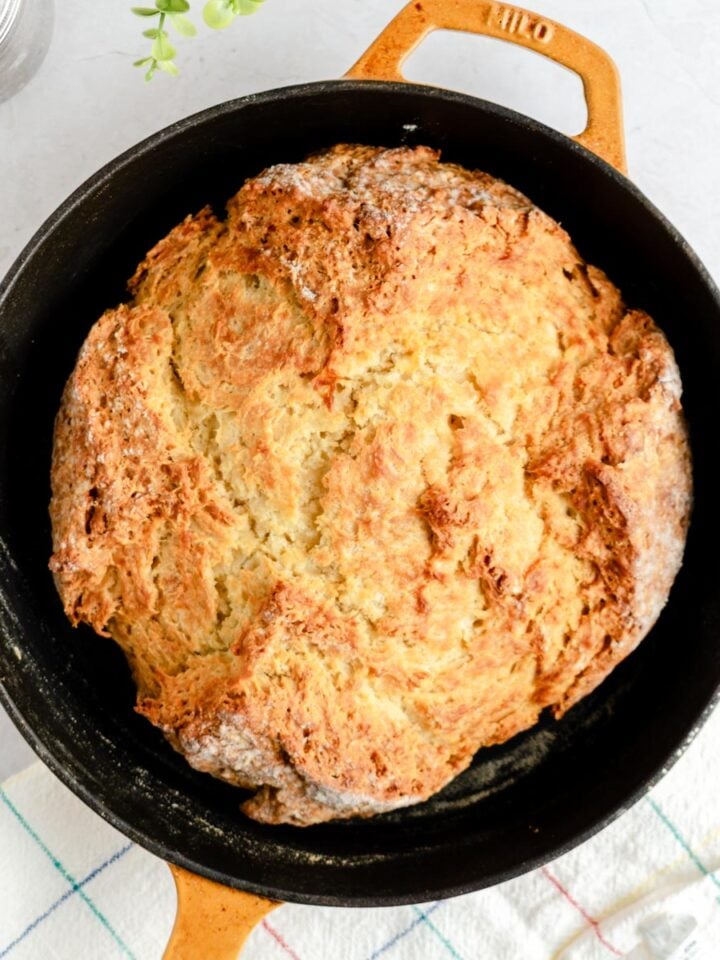 A round loaf of golden-brown soda bread sits in a black cast iron skillet with a tan handle, placed on a white and blue checkered towel, on a light-colored surface.