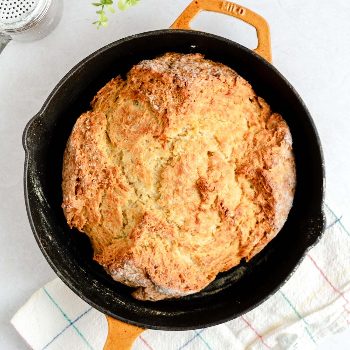 A round loaf of golden-brown soda bread sits in a black cast iron skillet with a tan handle, placed on a white and blue checkered towel, on a light-colored surface.