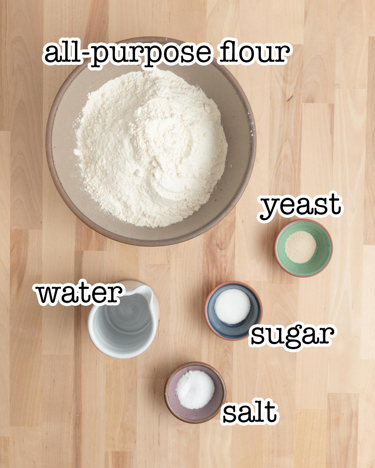 A bowl of all-purpose flour, a small bowl of yeast, a cup of water, and small bowls of sugar and salt are arranged on a wooden surface.