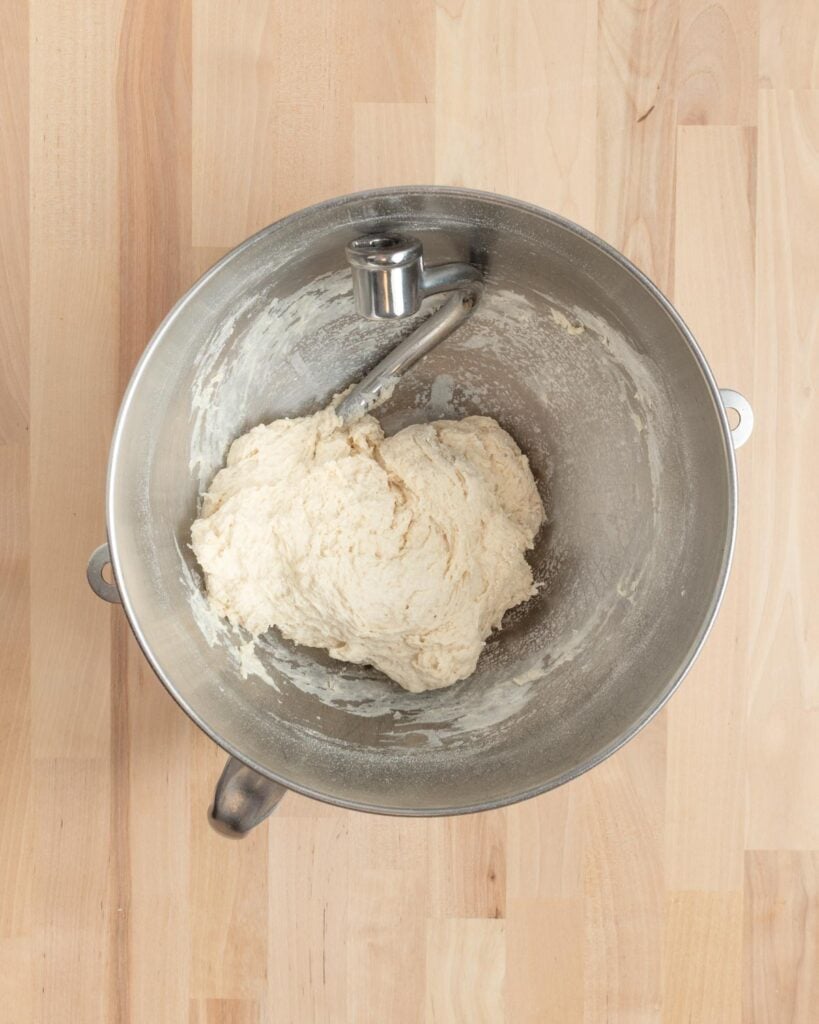 A metal mixing bowl with a dough hook attachment contains a rough and shaggy ball of dough. The bowl is placed on a light wooden surface.