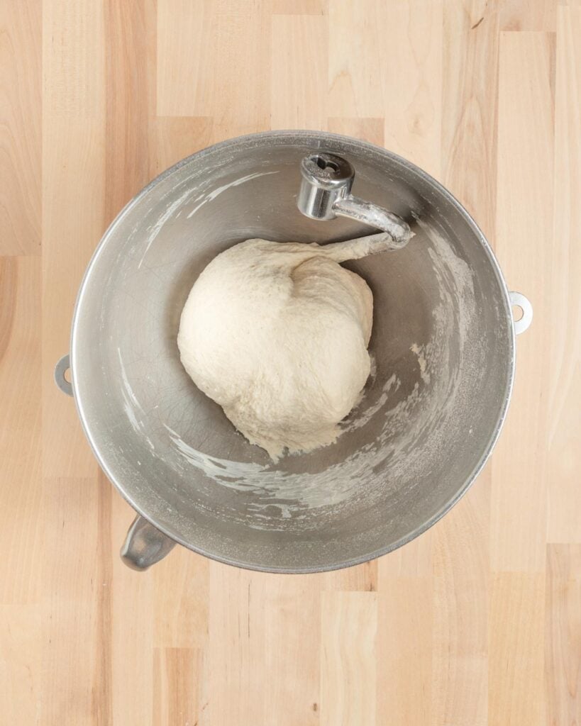 A smooth ball of bread dough sits in a metal mixing bowl with a dough hook attachment, on a light wooden surface. The dough is clinging slightly to the bowl around the edges but the walls of the bowl are mostly clear of dough and flour.