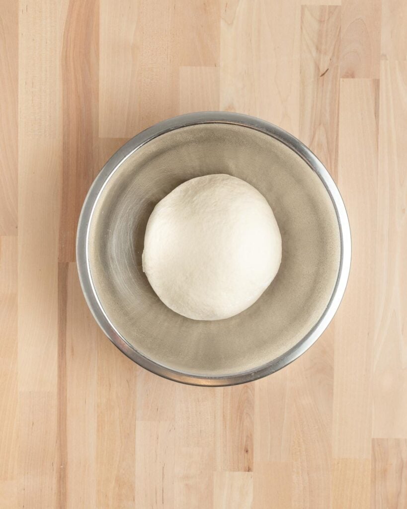 A round ball of dough sits in a large metal mixing bowl on a light wooden surface, viewed from above.