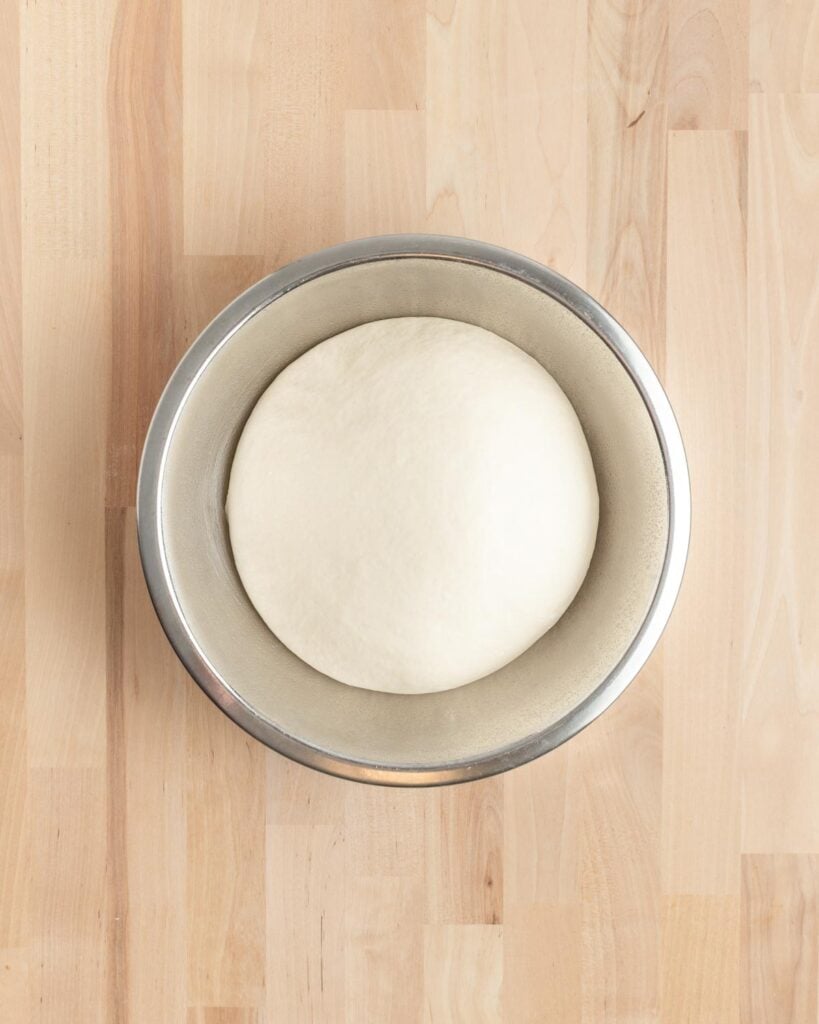 A round, smooth ball of white dough rising in a stainless steel bowl, placed on a light wood surface.