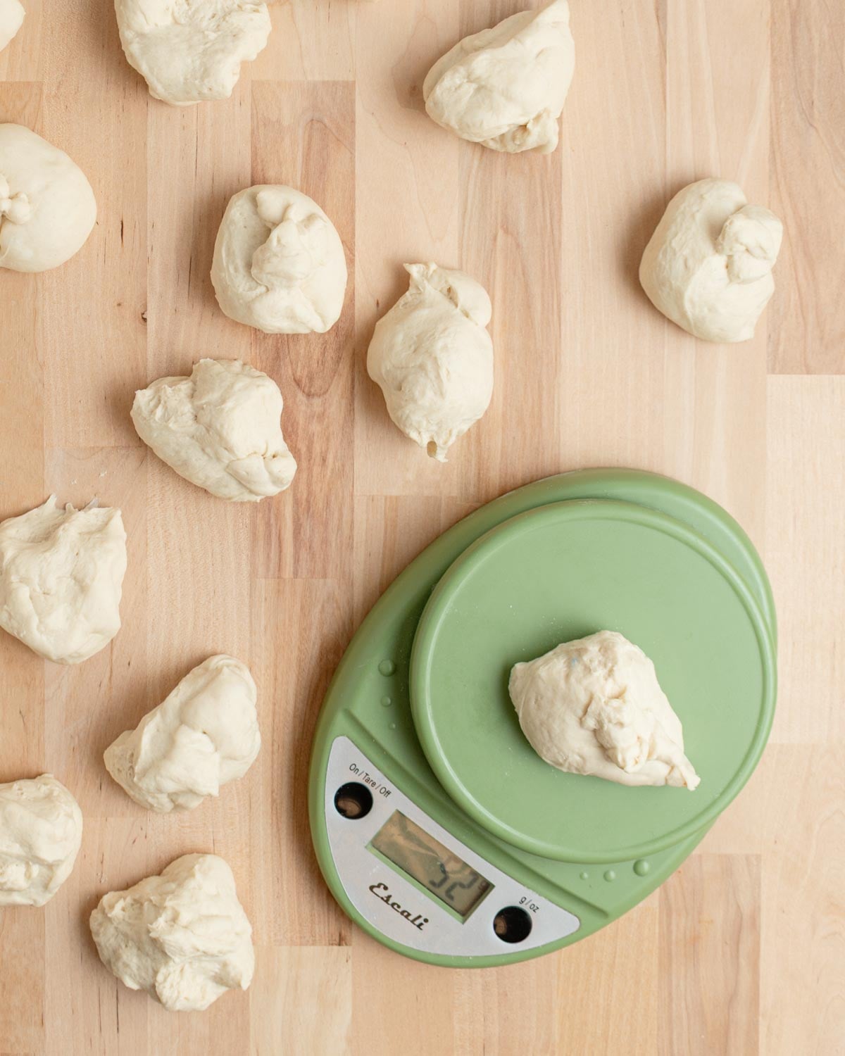Several pieces of dough are placed on a light wooden surface. One dough piece is being weighed on a green digital kitchen scale, which displays 27.1 grams.